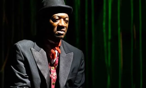A man in a dark suit and hat sitting against a green curtain background.