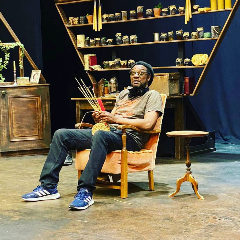 Man sitting in a vintage armchair holding incense sticks, wearing glasses, a beanie, and casual clothes, in a room with dark walls and shelves filled with jars and books.