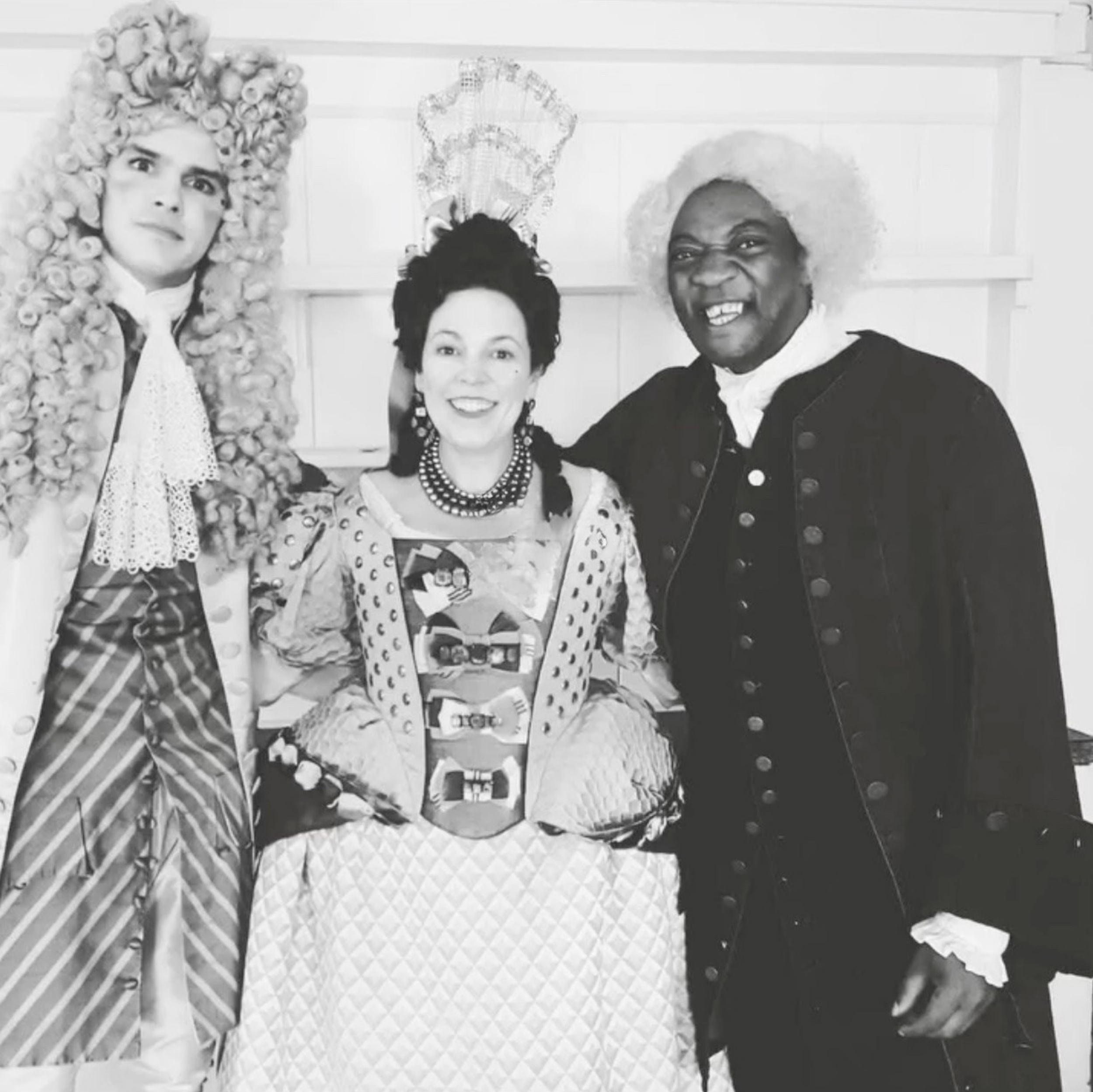 Four people dressed in colonial costumes, including wigs and period clothing, posing together for a photo in front of a plain wall.