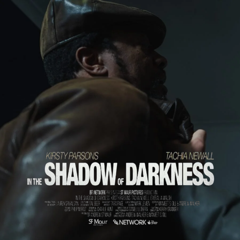 A man wearing a brown leather jacket and a brown cap, facing sideways, in a dimly lit setting. The movie poster for "In the Shadow of Darkness" is displayed, featuring the names Kirsty Parsons and Tacha Newall.
