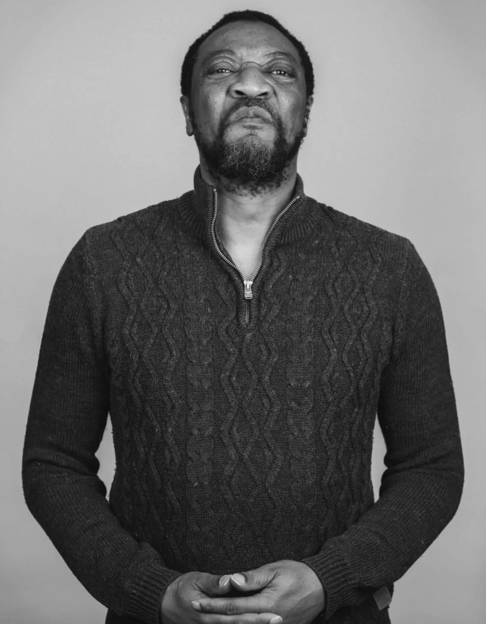 Black and white photo of a man with a beard and mustache, wearing a dark, textured sweater with a zipper, crossing his hands in front of him, standing against a plain background.