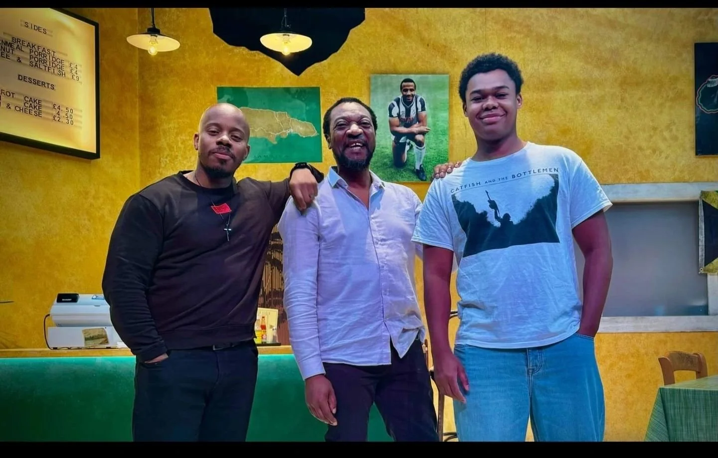 Three men standing together in a restaurant with yellow walls and framed photos, smiling at the camera.