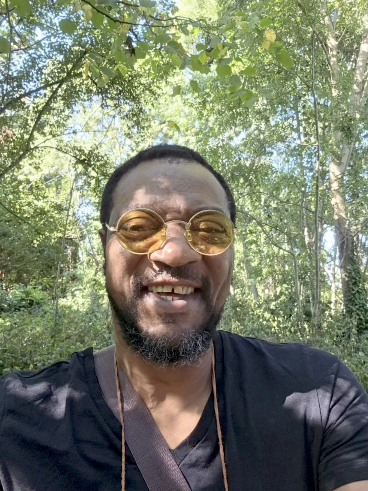 A man with glasses smiling outdoors in a forested area, wearing a black shirt and a brown strap across his chest.