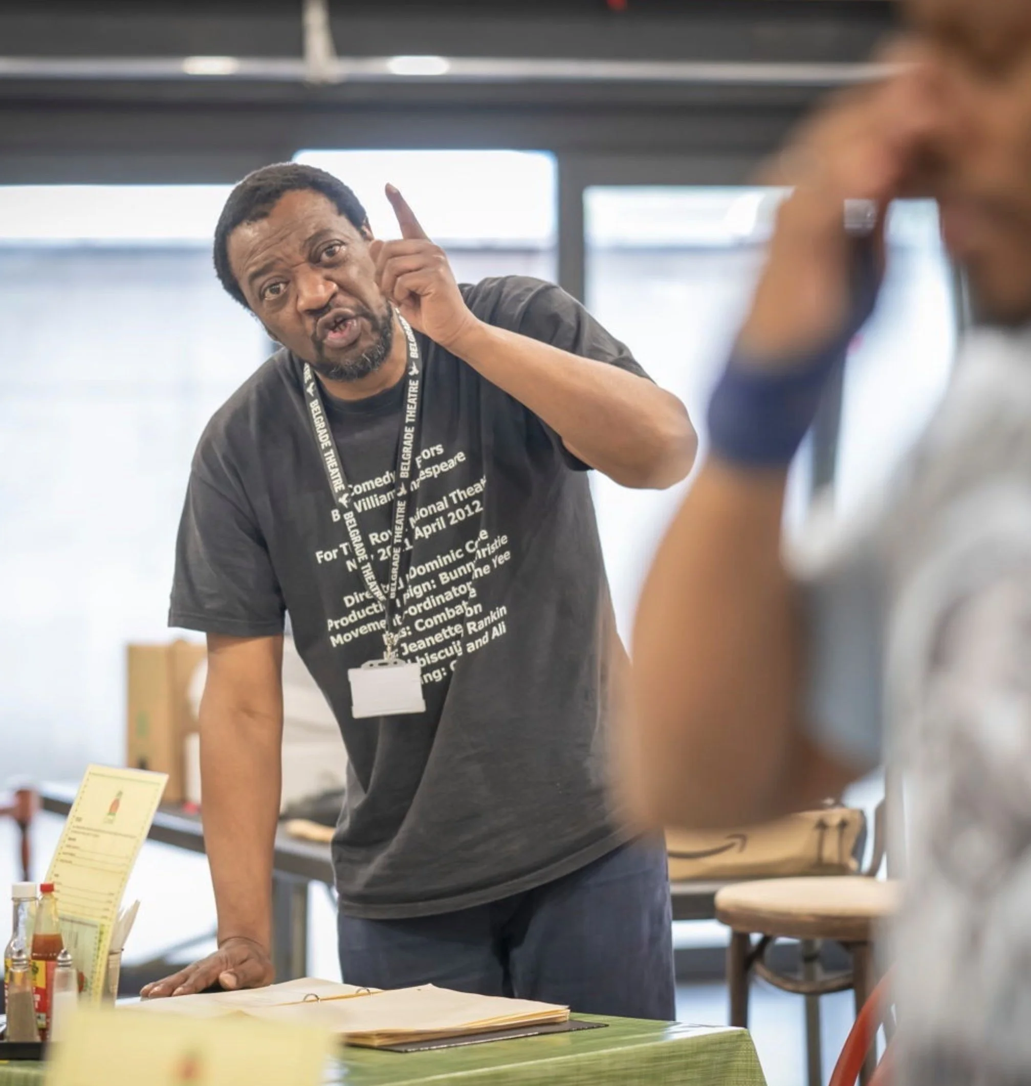 Man with short black hair, beard, wearing a black shirt with white text, gesturing with his right hand near his ear while speaking to a person on the right in a room with large windows and tables.