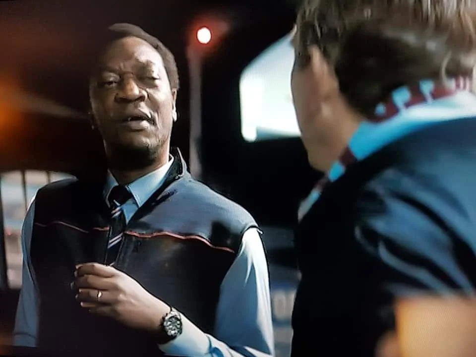 A scene from a movie or TV show featuring two men in conversation inside a vehicle at night, with one appearing to be explaining or emphasizing a point.