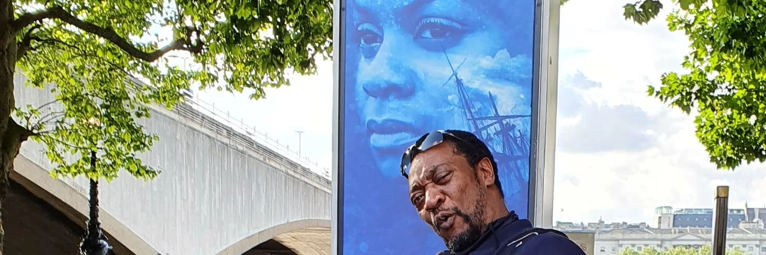 A man with dark hair and beard making a facial expression in front of a large blue billboard with a woman's face painted on it and a ship outline, outdoors on a sunny day with green trees.