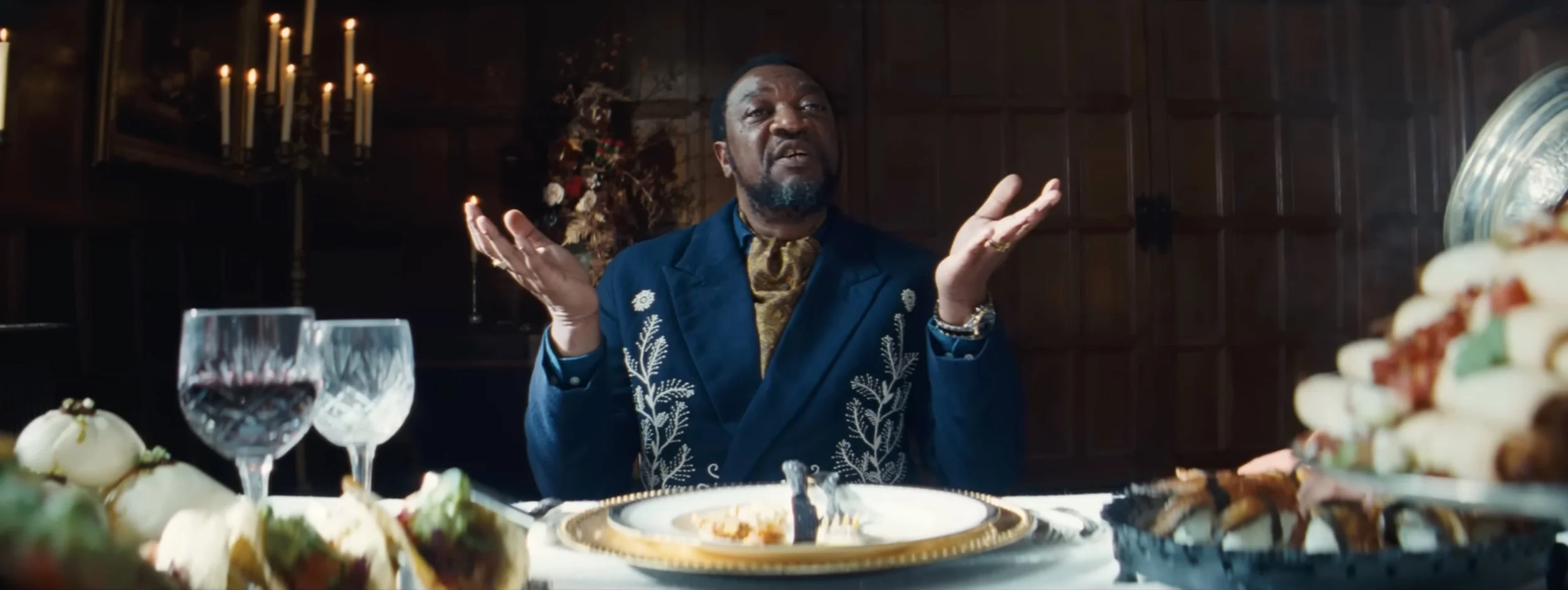 A man sitting at a dining table with food, wine glasses, and plates, gesturing with his hands and speaking, in a festive setting with candles and a Christmas decoration in the background.