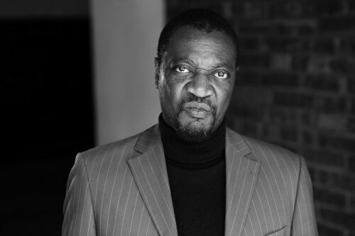 Black man in a striped suit with a black turtleneck, standing against a brick wall background.