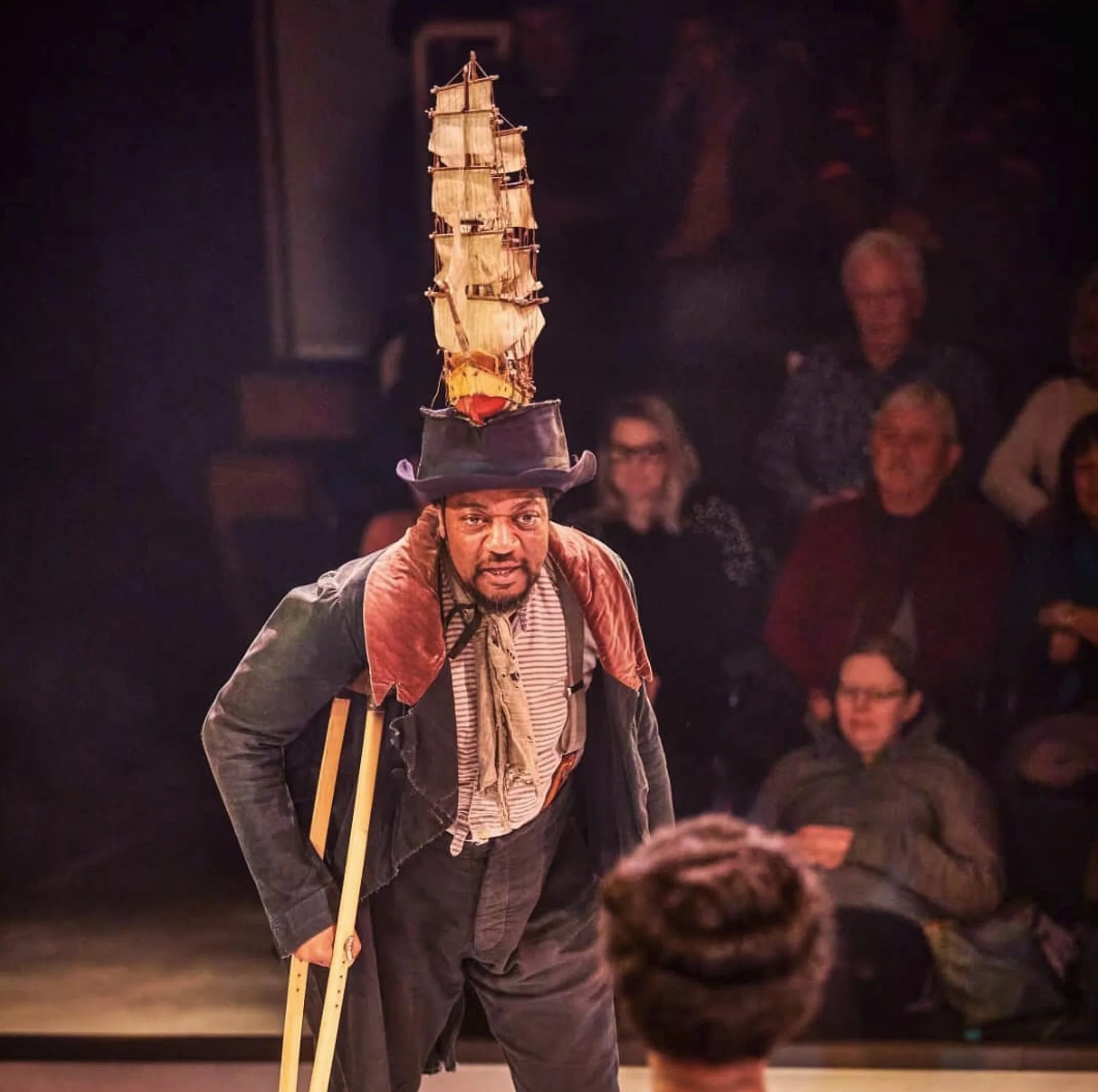 A man performing a balancing act with a small model ship on his head during a show, with an audience watching in the background.