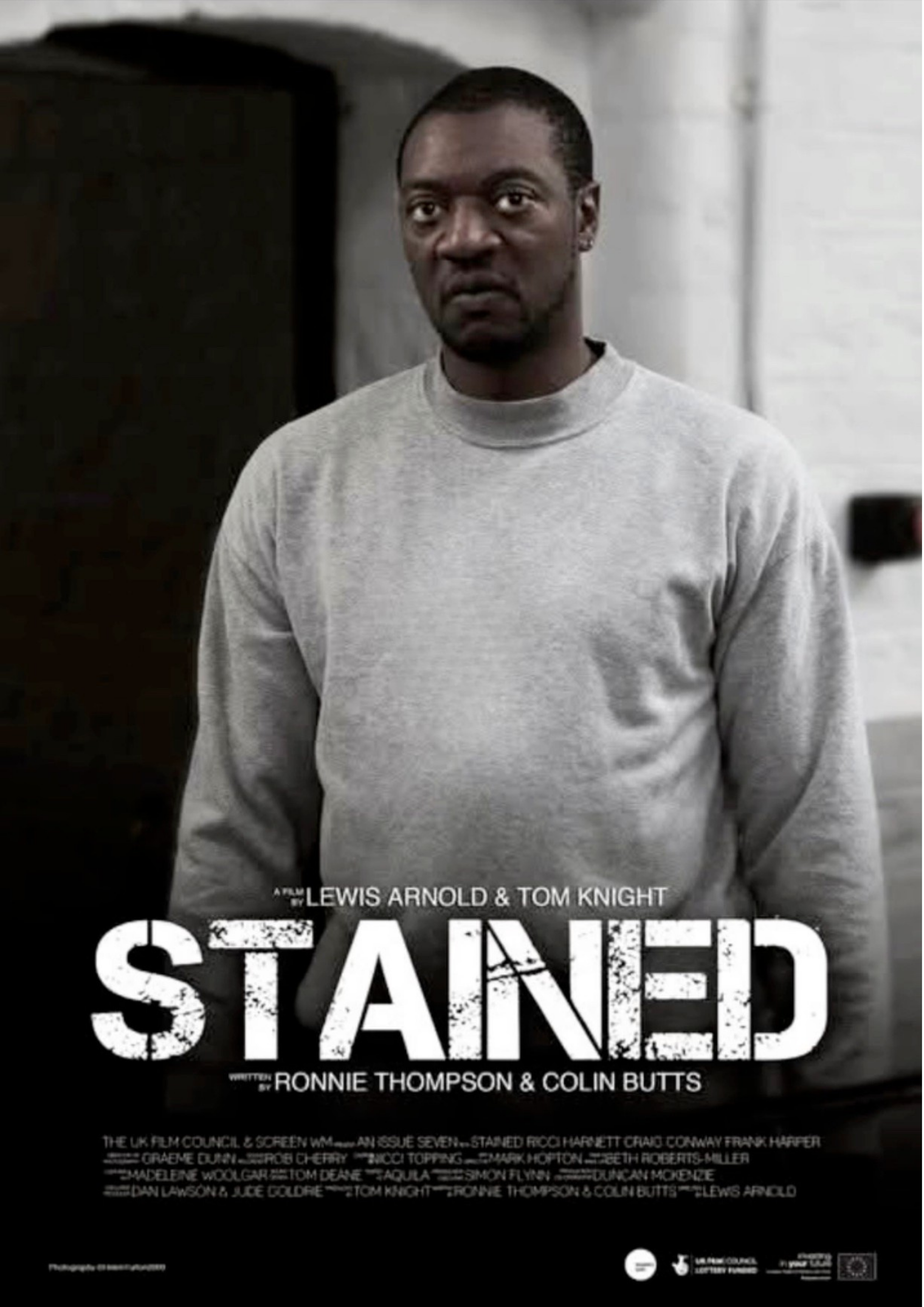 A man with short hair and a serious expression wearing a light gray sweatshirt, standing indoors with a white brick wall in the background, with a film poster visible in the foreground.