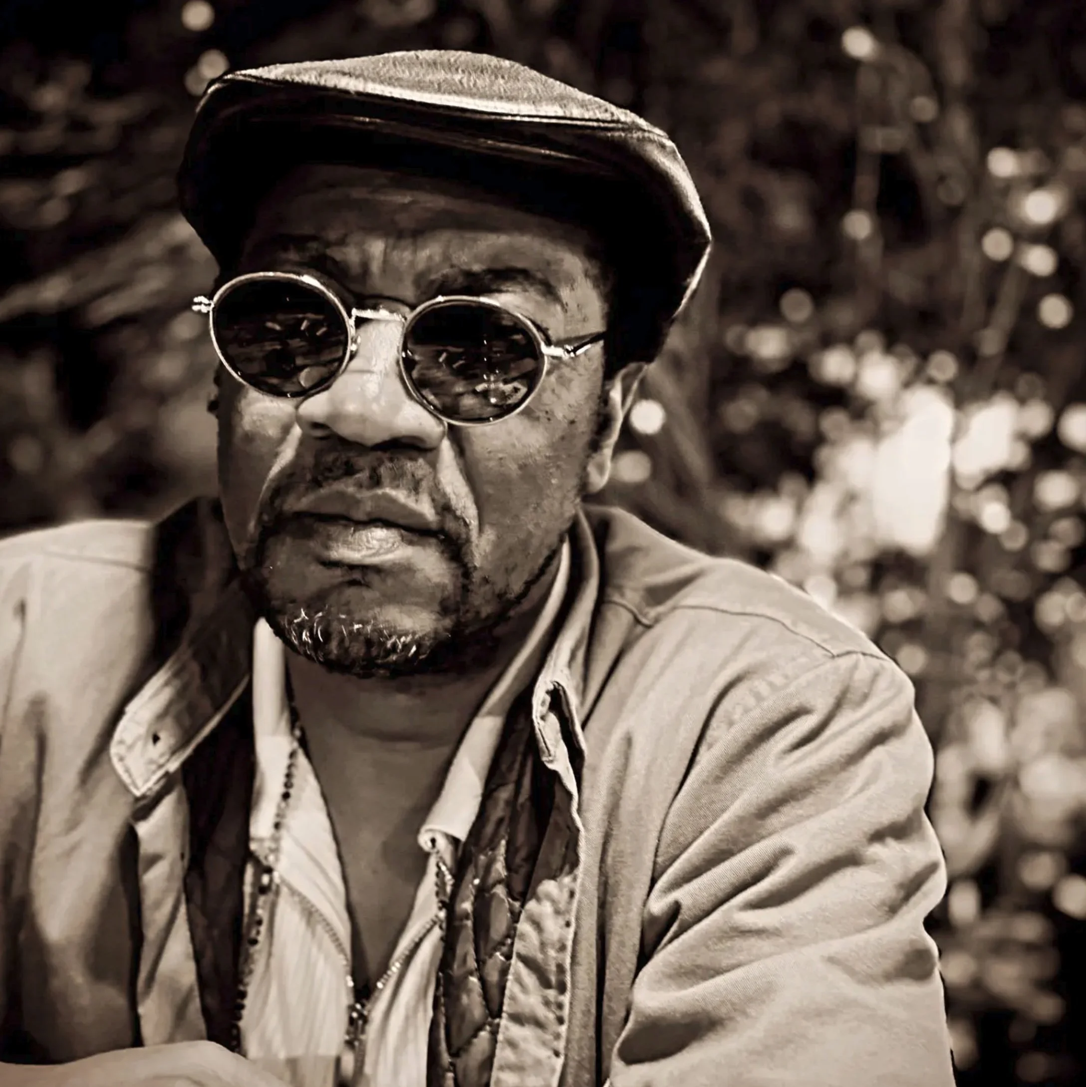A man wearing round sunglasses, a fedora hat, and a light-colored jacket with a zipper, with blurred background suggesting an outdoor setting, possibly in sunlight.