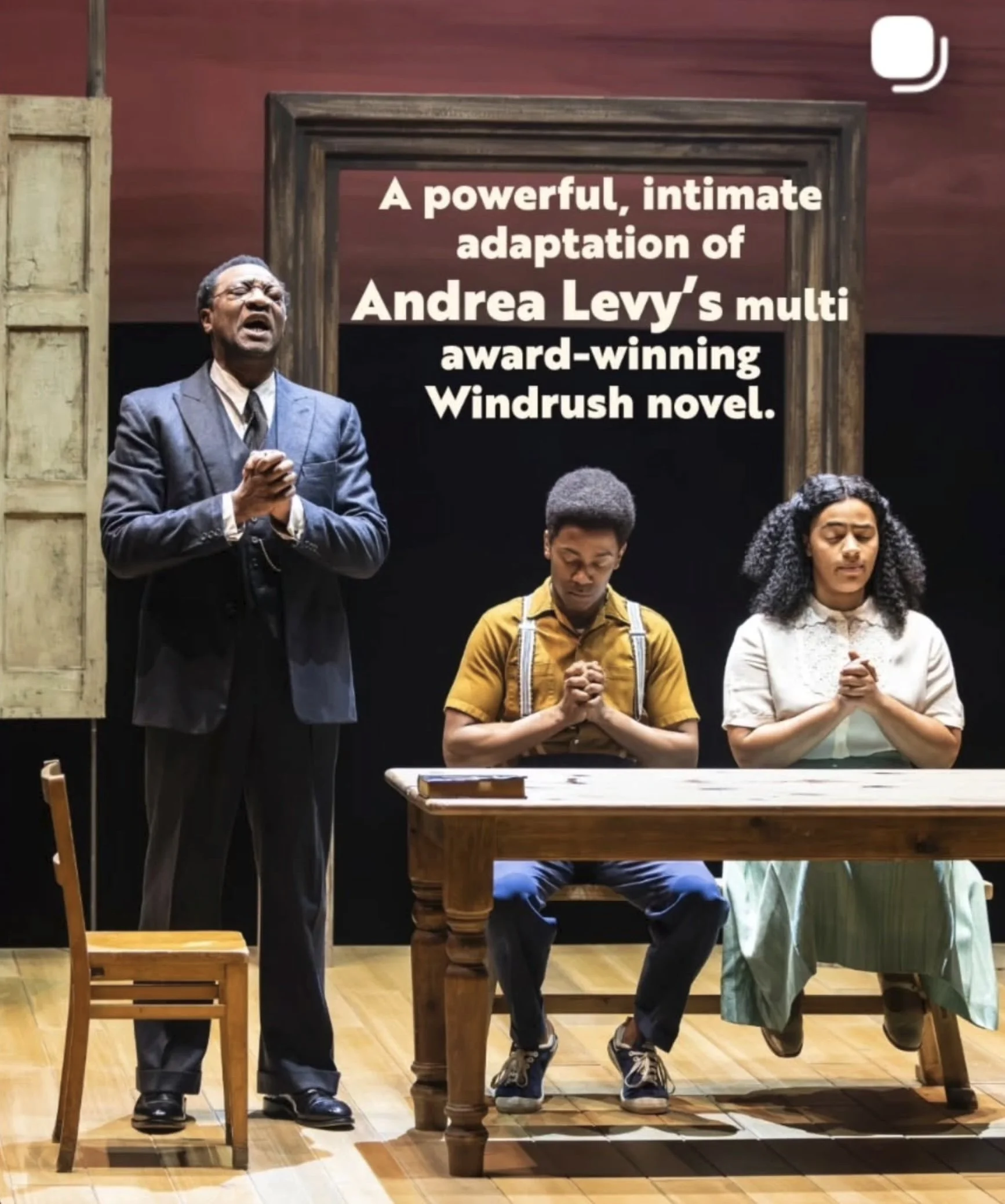 A group of four people praying with heads bowed and hands clasped in front of a wooden table and chairs in a room with a red wall. The background features a photo frame with a dark background and a piece of furniture with a book on it.