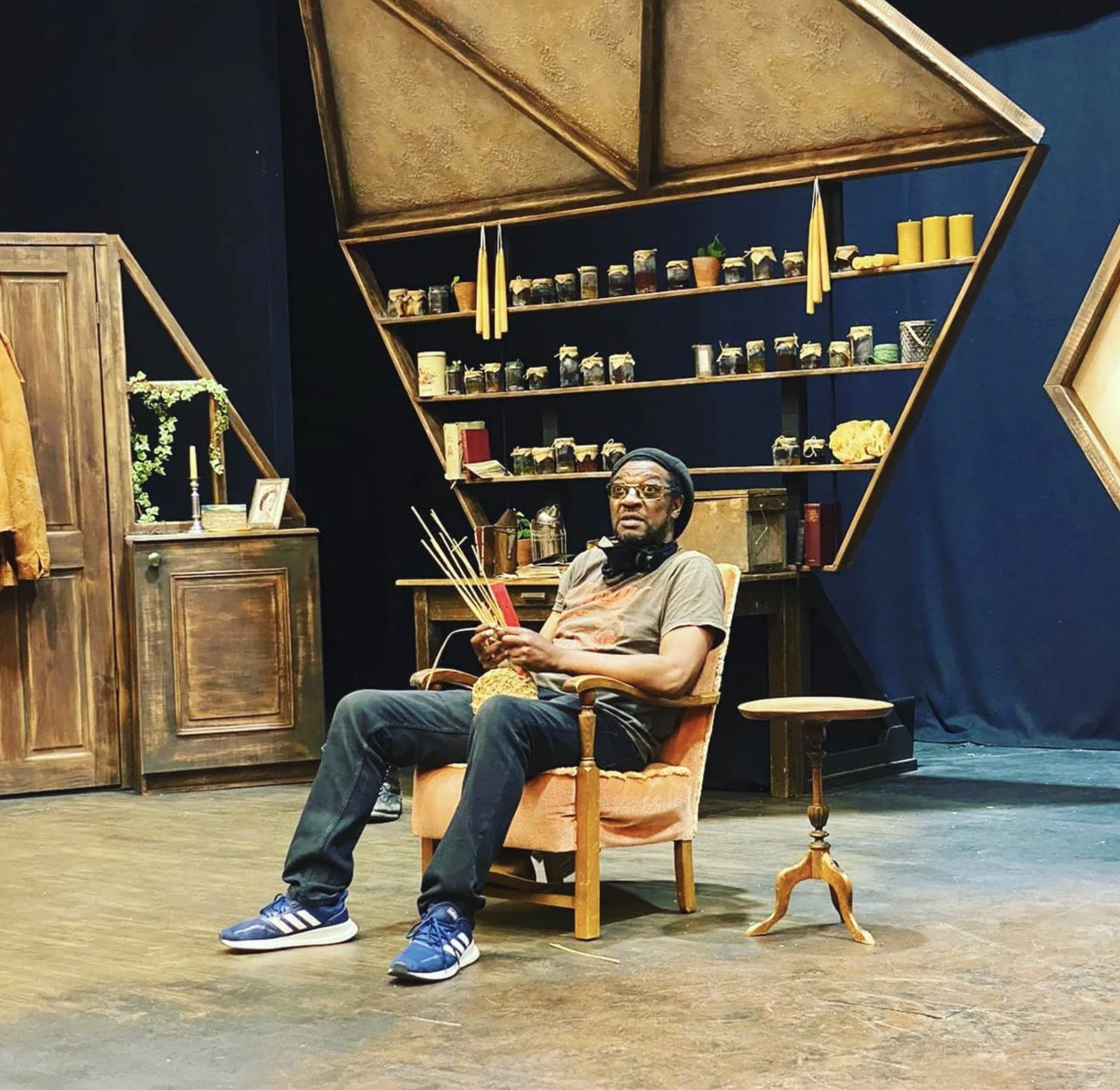 A man sitting in a wooden chair on a stage, holding knitting needles and yarn, with bookshelves and decorative items behind him.