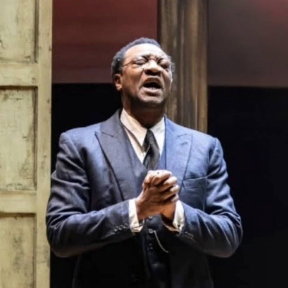 An old man in a suit with glasses, eyes closed, praying with clasped hands.