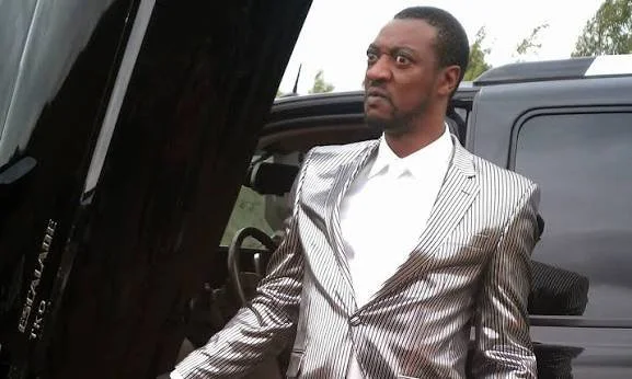 A man in a silver pinstripe suit standing next to a black vehicle, with an intense, angry facial expression.