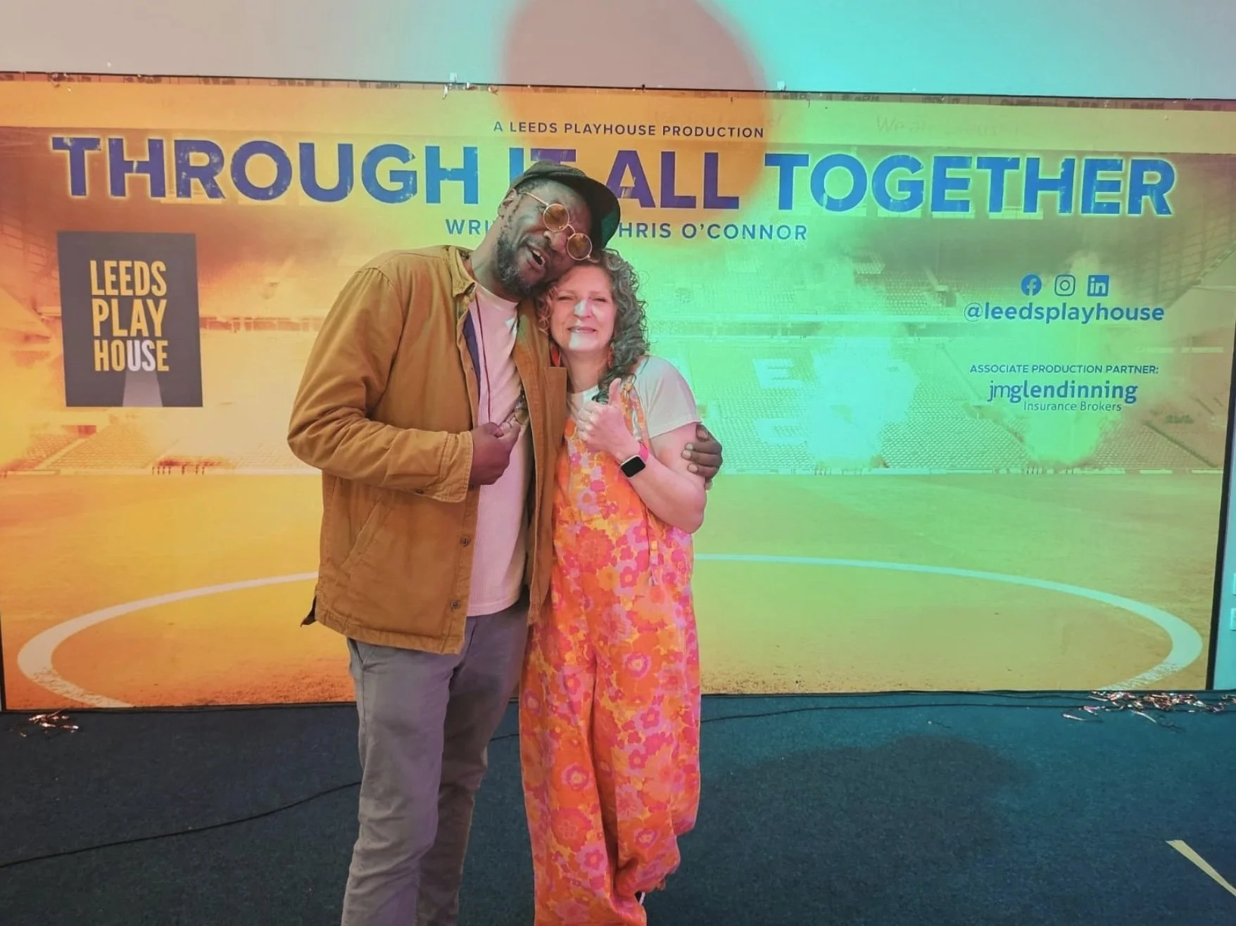 Two people, a man wearing glasses and a woman with curly hair, are smiling and standing together in front of a colorful backdrop promoting a Leeds Playhouse production titled "Through It All Together." The man is giving a thumbs-up, and the woman is 