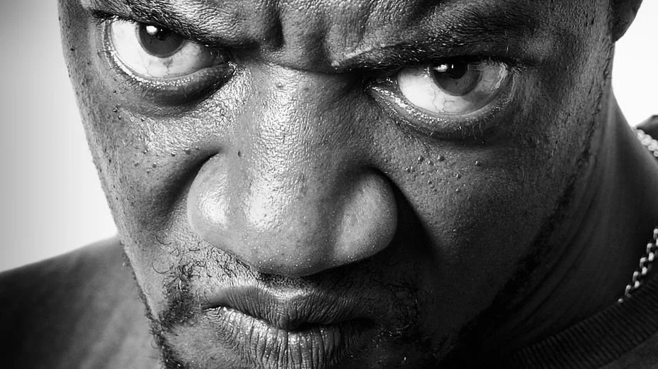 Close-up black and white photo of a man's intense facial expression with piercing eyes and furrowed brows.