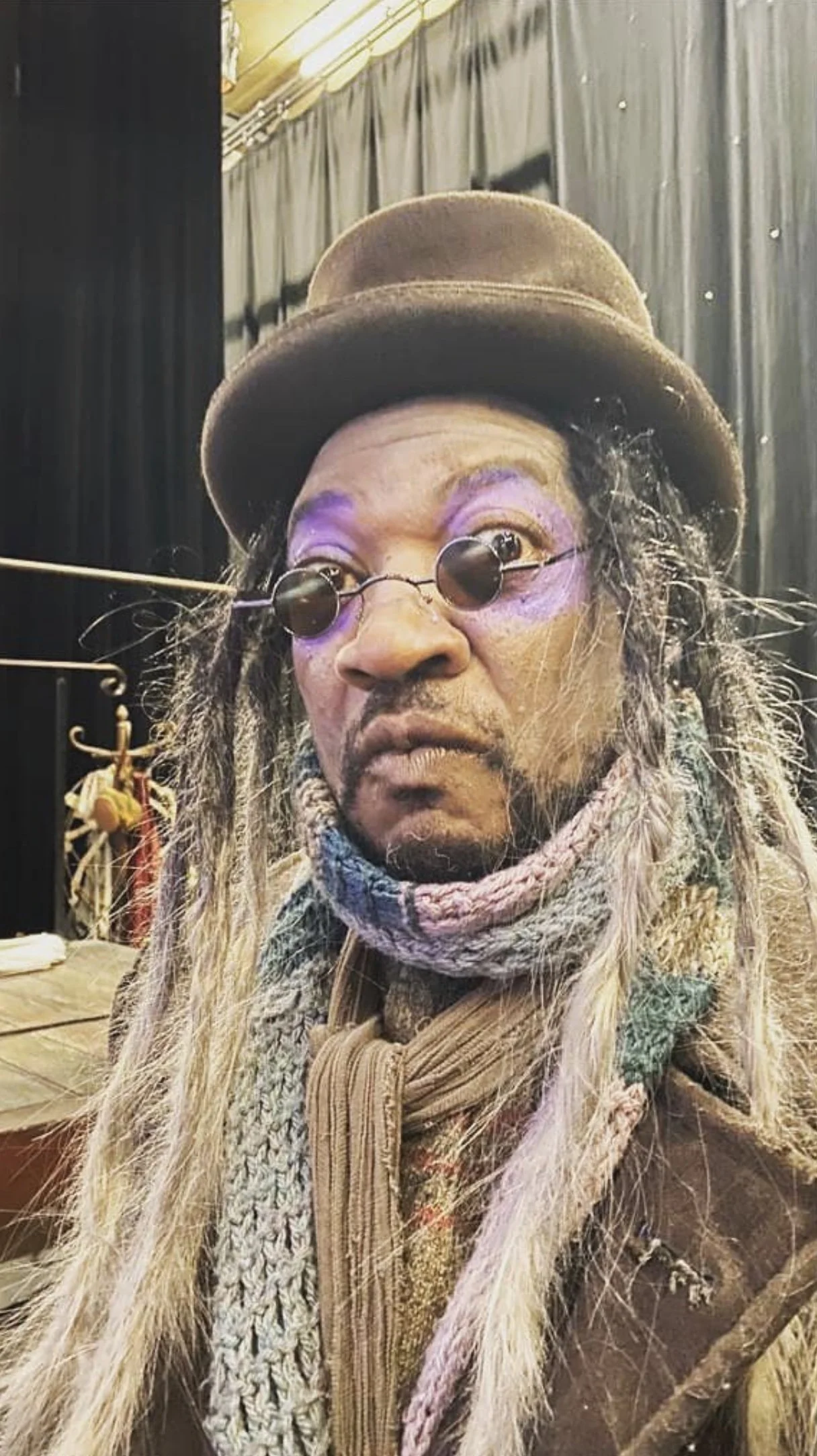 A person with long, blonde dreadlocks wearing a brown hat, round sunglasses, purple eye makeup, and a multicolored scarf and coat inside a room with black curtains and a table in the background.