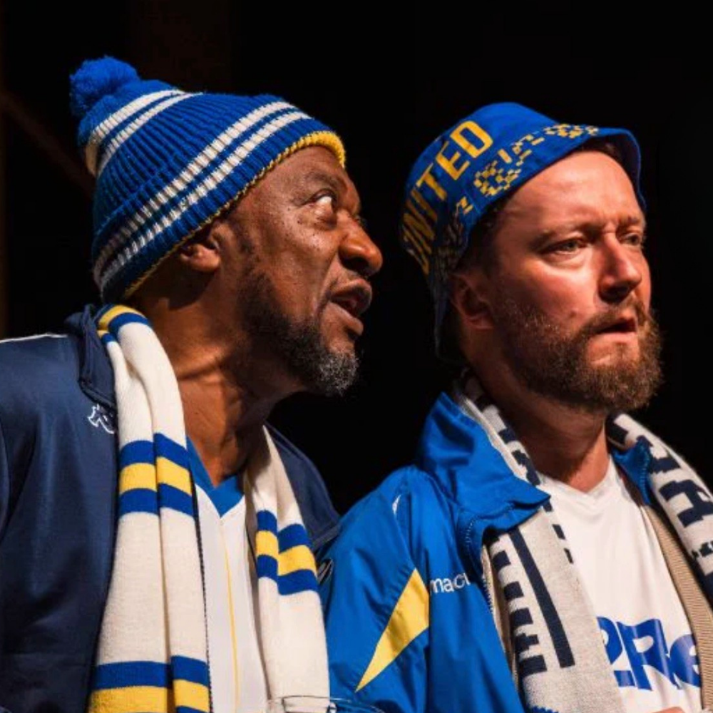 Two men wearing blue and yellow scarves and beanies, standing close together against a black background, with serious expressions.