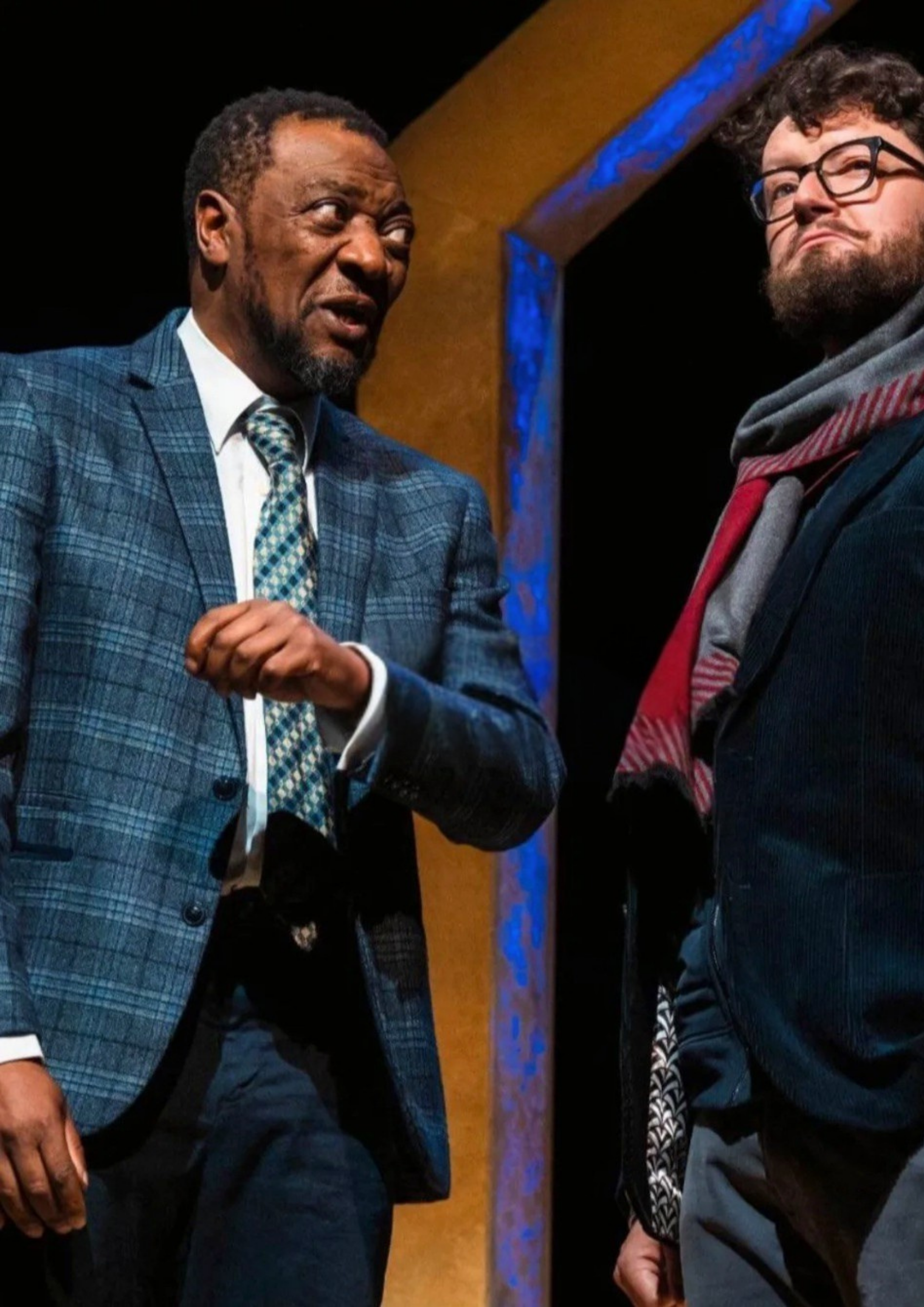 Two men engaged in conversation, one wearing a blue checkered suit and tie, the other a dark jacket with a red scarf, in a setting with a dark background and a colorful arch.