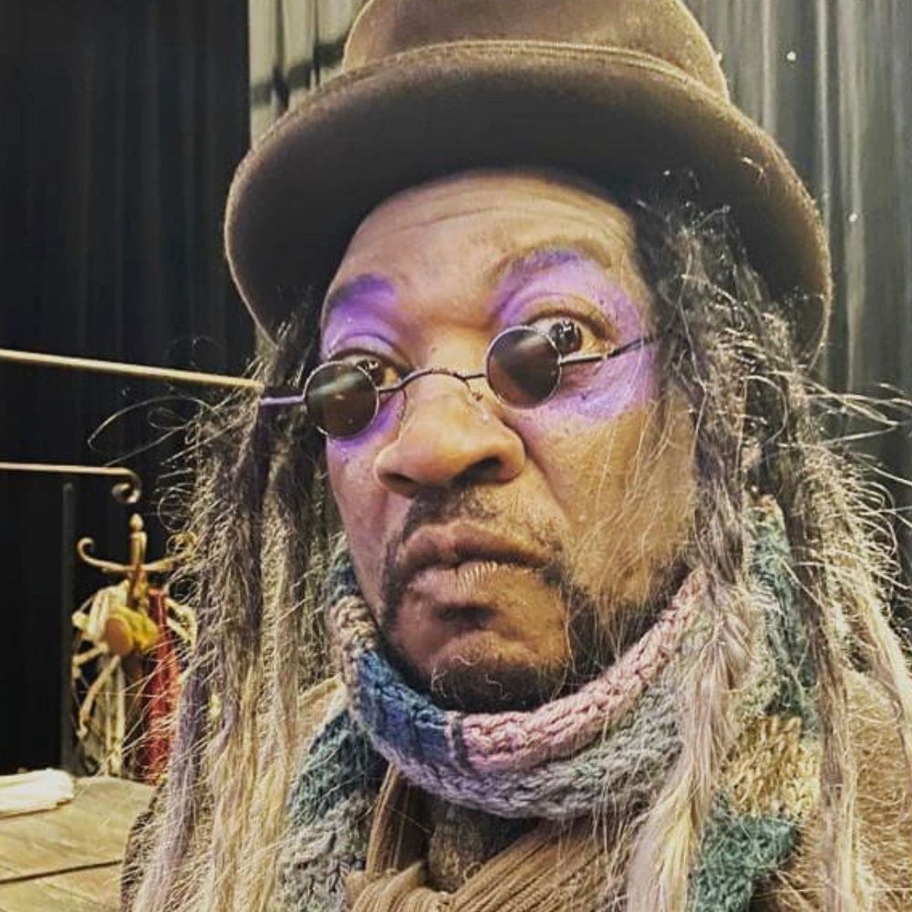 A person with dreadlocks wearing a brown hat, round sunglasses, and a multicolored knitted scarf, with purple eyeshadow and a surprised expression.