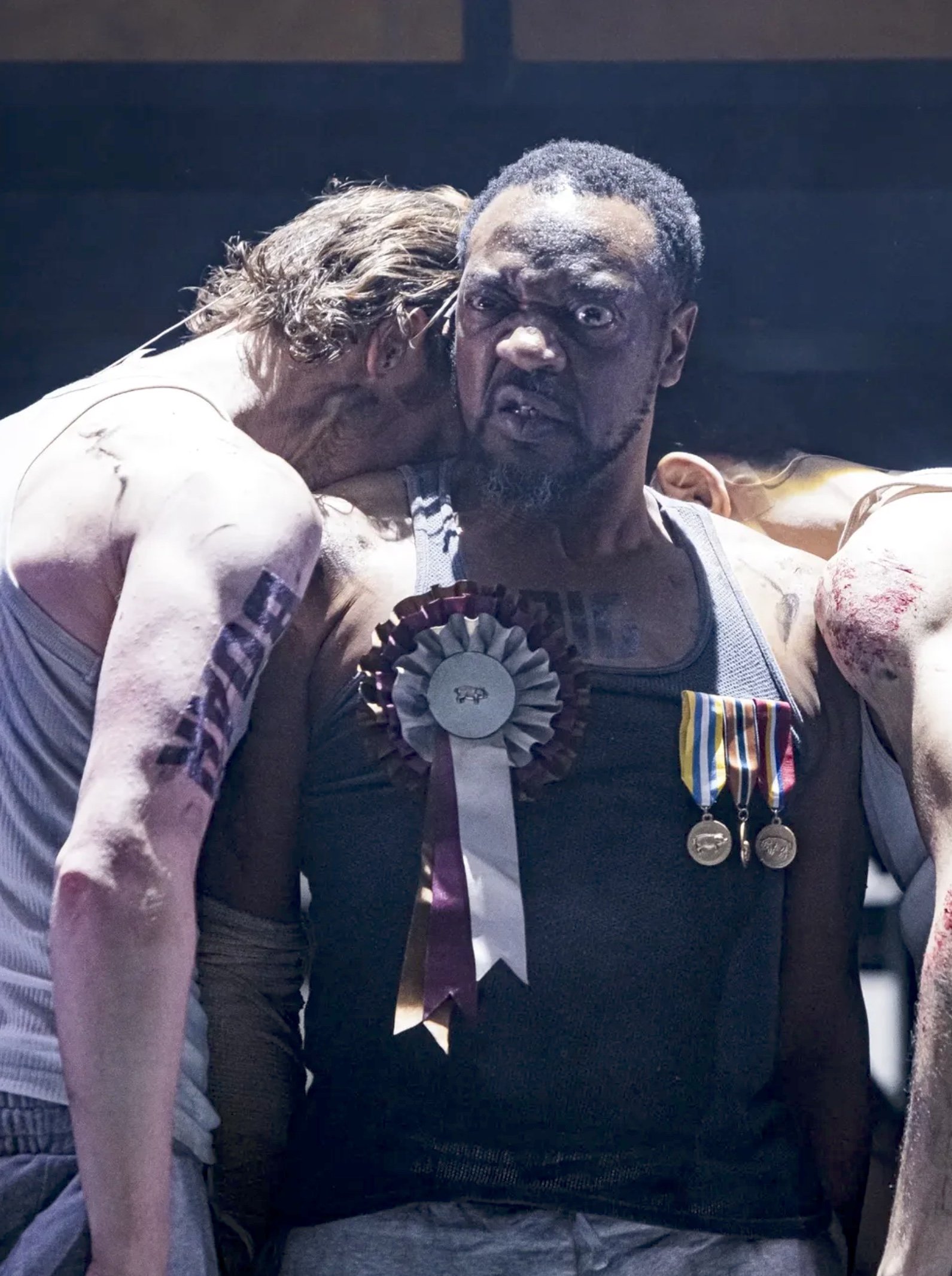 Two men, one with long blond hair and the other with short gray hair and a beard, sitting close together. The man with gray hair is wearing medals and a ribbon, looking directly at the camera with a serious expression. The other man is leaning in wit