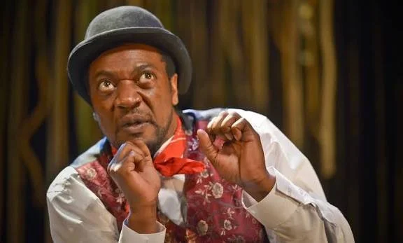 A man dressed as a performer or actor with a gray hat, a red bow tie, and a patterned vest, making a humorous face with his hands near his face in a theatrical setting.