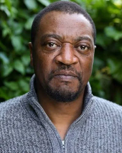 Close-up portrait of a man with dark skin, short curly hair, and a beard, wearing a gray zip-up sweater, standing outdoors with green foliage in the background.