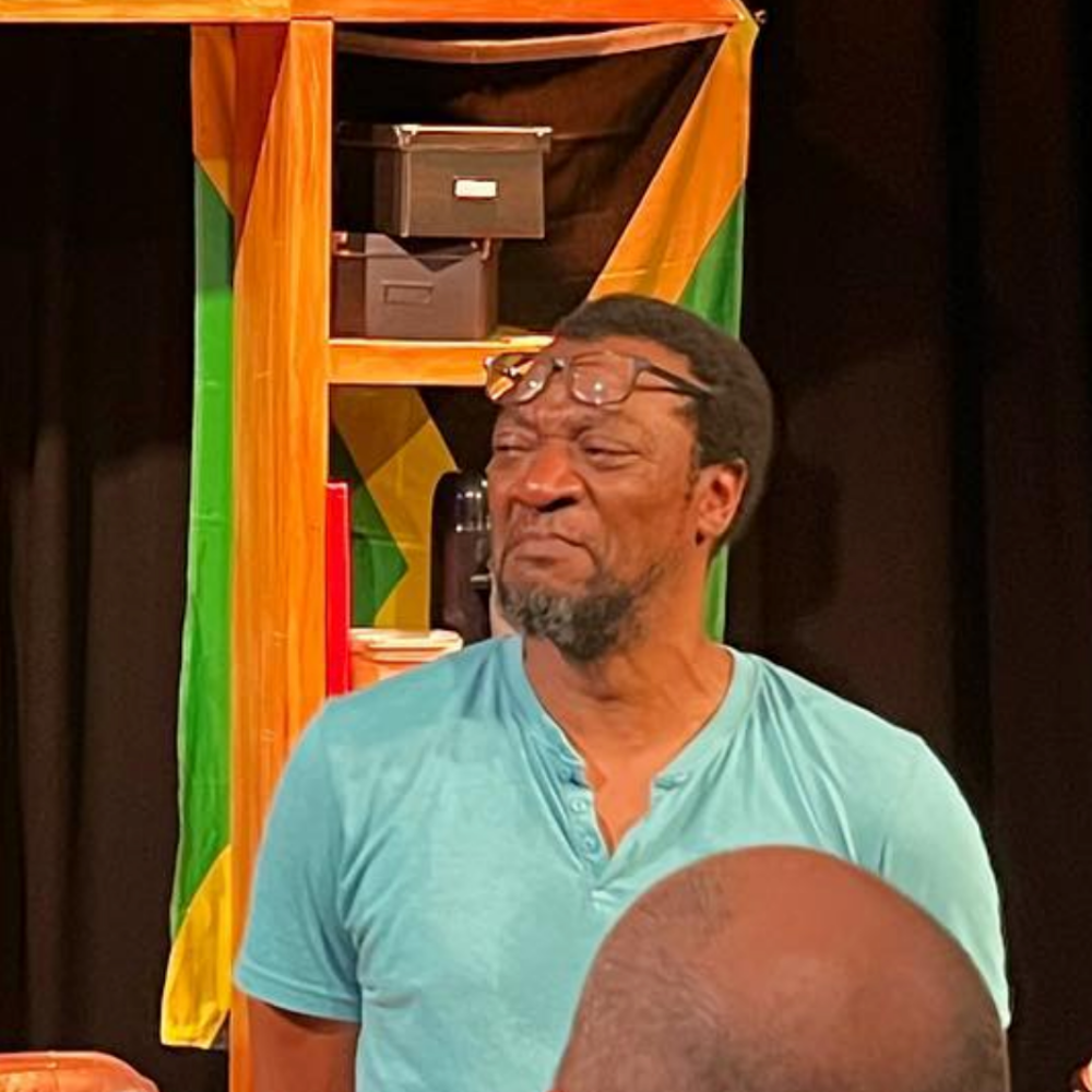 A man with glasses on his forehead making a facial expression with his eyes squinted and mouth slightly pursed, standing in front of a colorful background with green, yellow, and orange patterns.