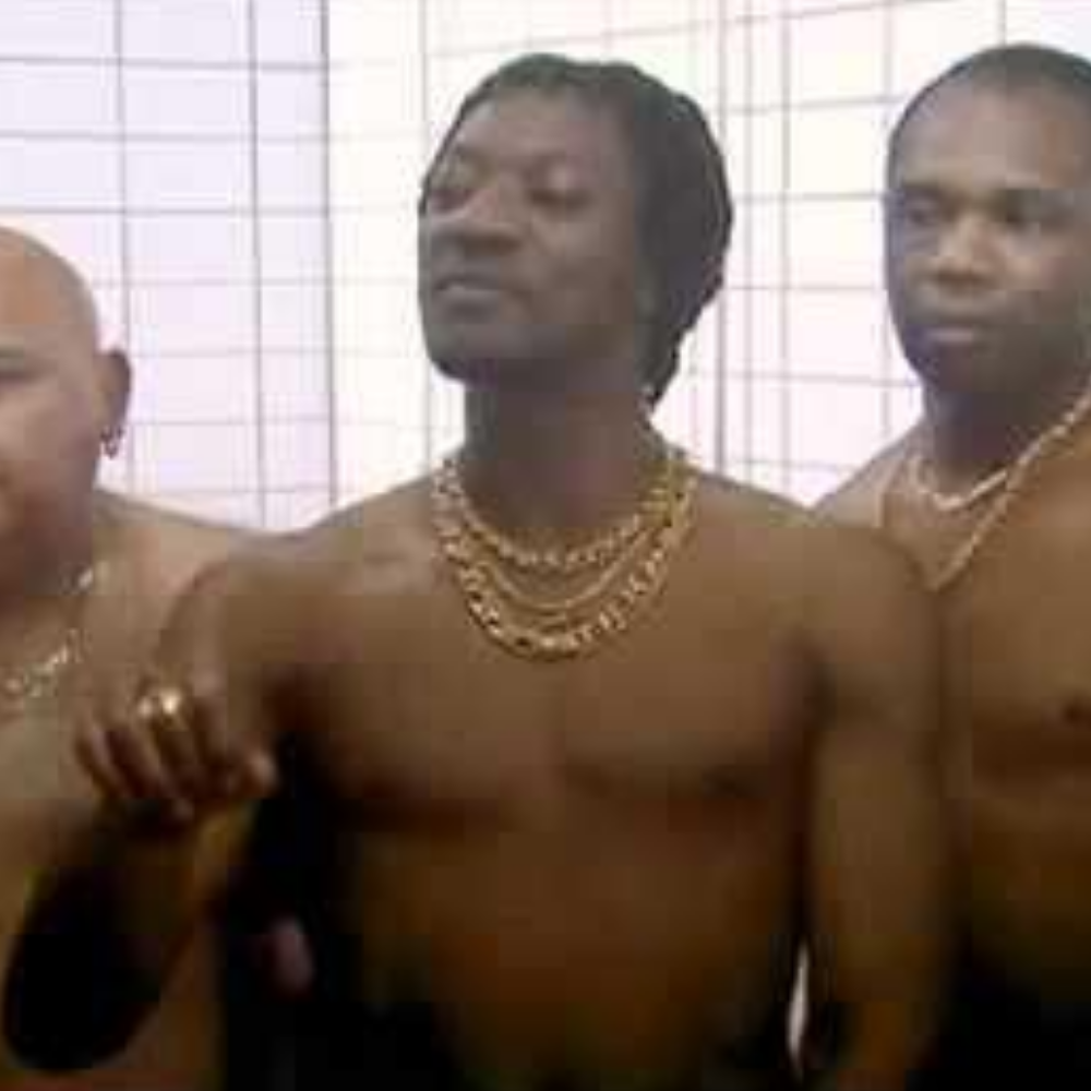 A group of shirtless men wearing gold chains in front of a wire mesh background.