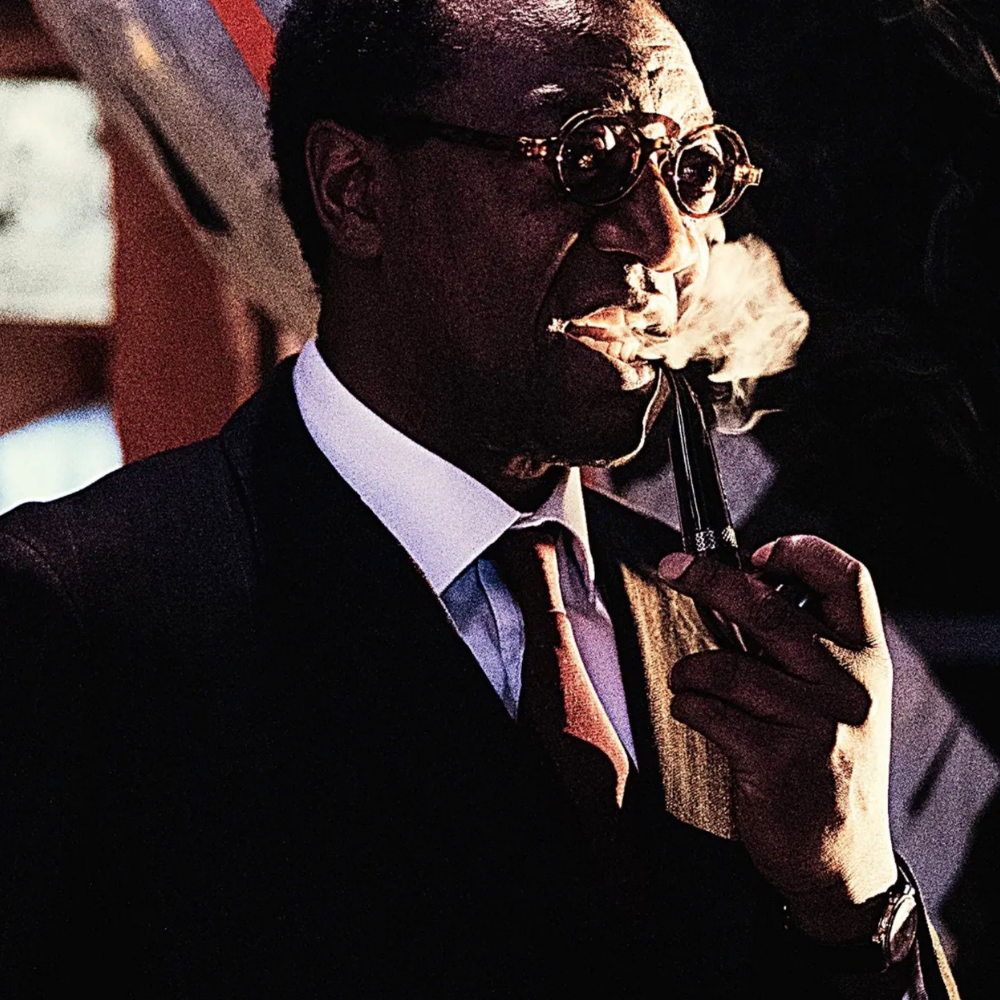 A man in a suit and glasses is smoking a pipe, with smoke coming out of his mouth.
