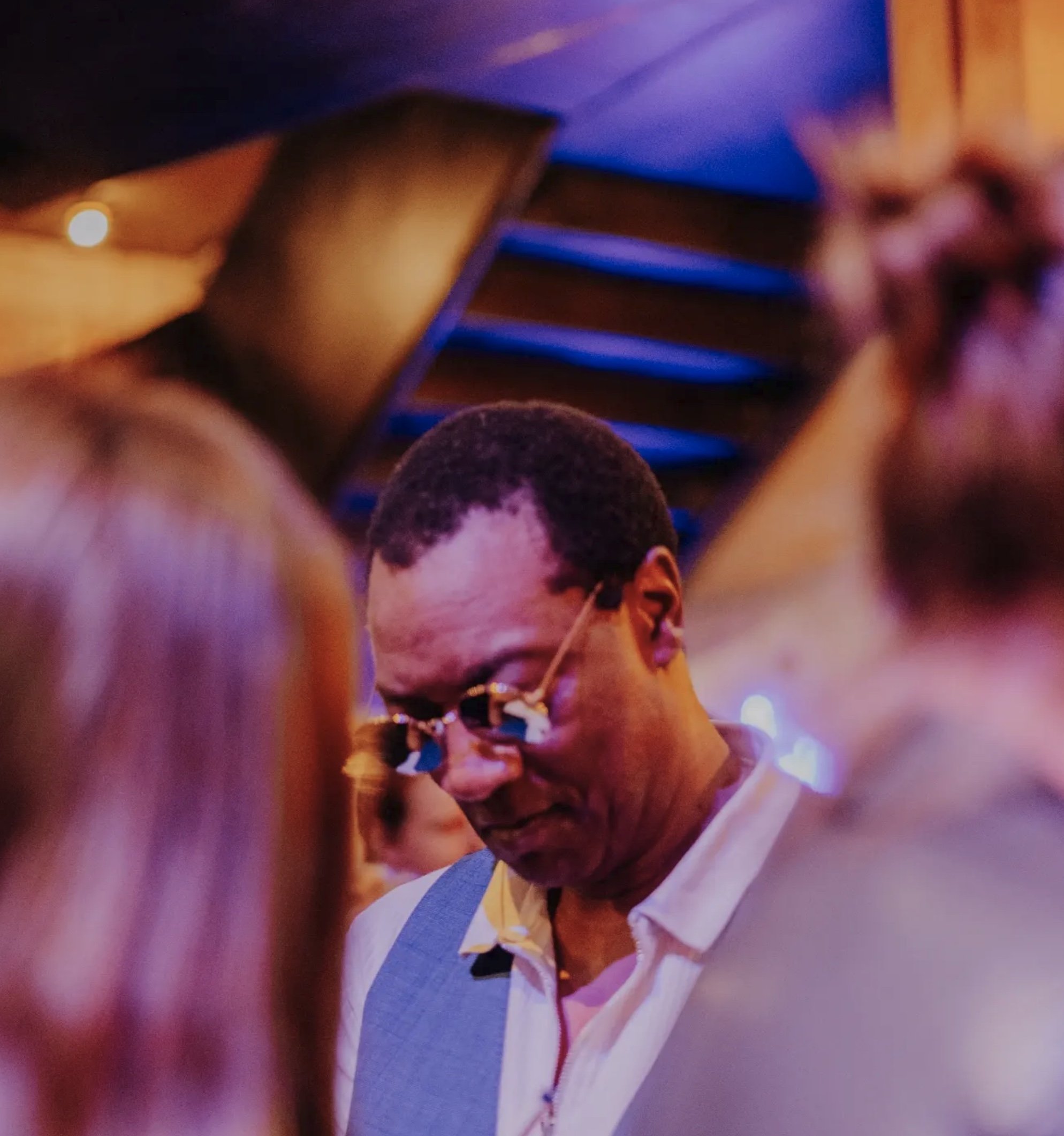 Close-up of cheering man at a concert or event wearing glasses, with blurred background and vibrant purple and pink lighting.