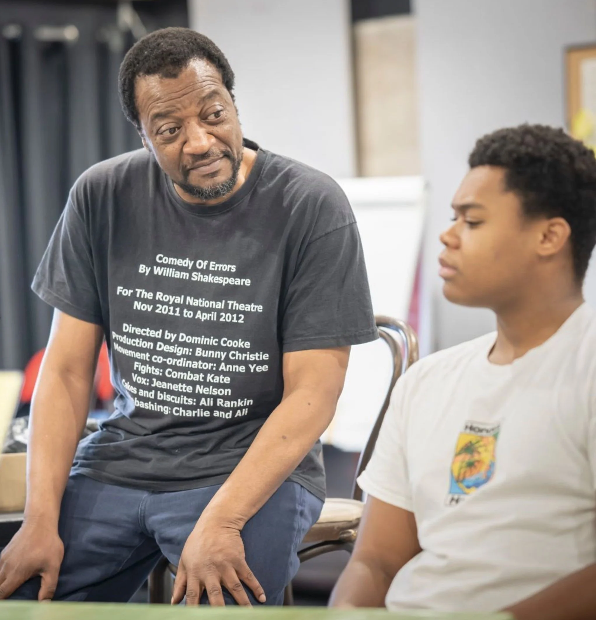 An older man with short dark hair and a beard, wearing a black T-shirt with white text, is talking to a young boy with short curly hair, who is wearing a white T-shirt with a colorful map print. They are seated in a room with bookshelves and a poster