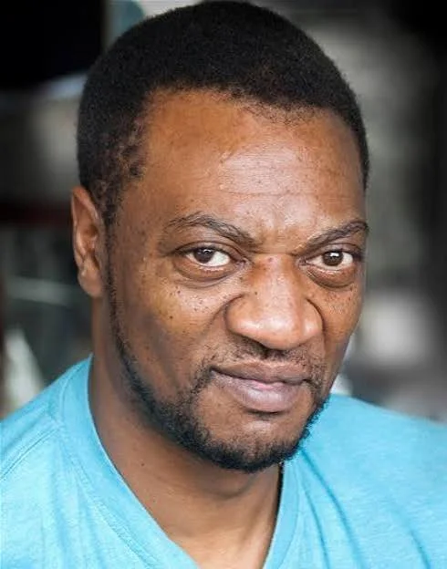 Close-up of a man with short black hair, beard, wearing a light blue shirt, looking at the camera.