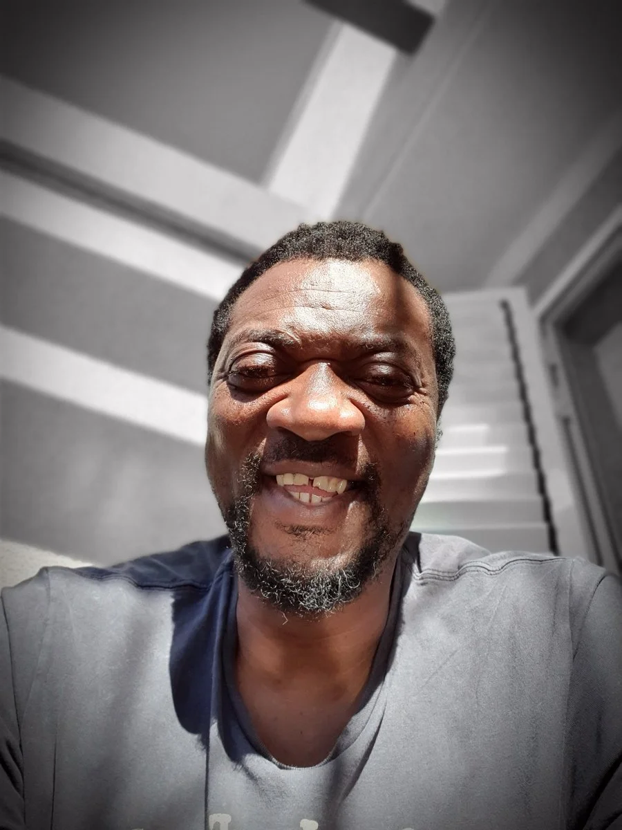 A smiling black man with facial hair taking a selfie in front of a background with stairs and a door.