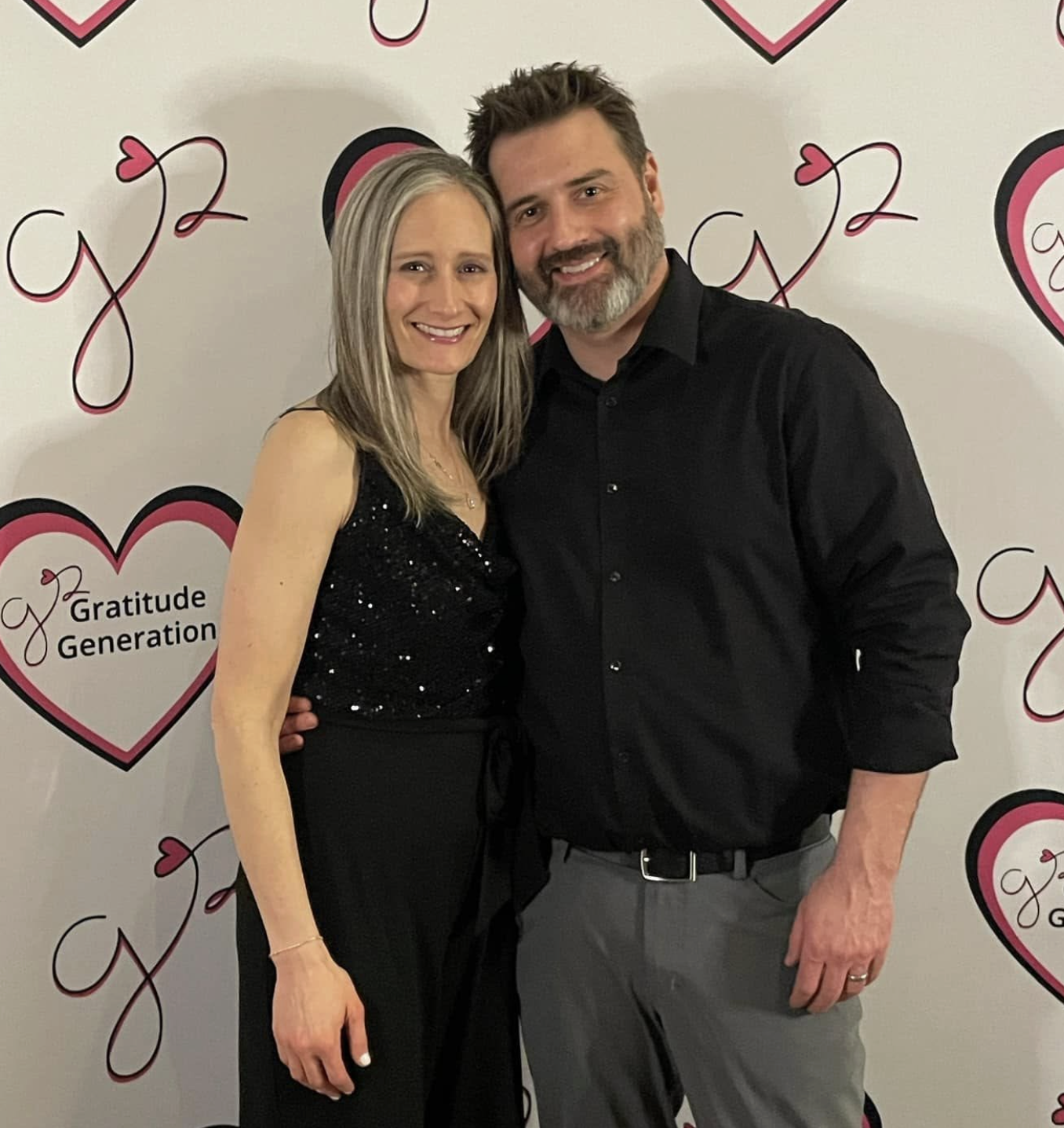 A woman and a man standing close together, smiling, in front of a backdrop with pink and black hearts and the words 'Gratitude Generation.'