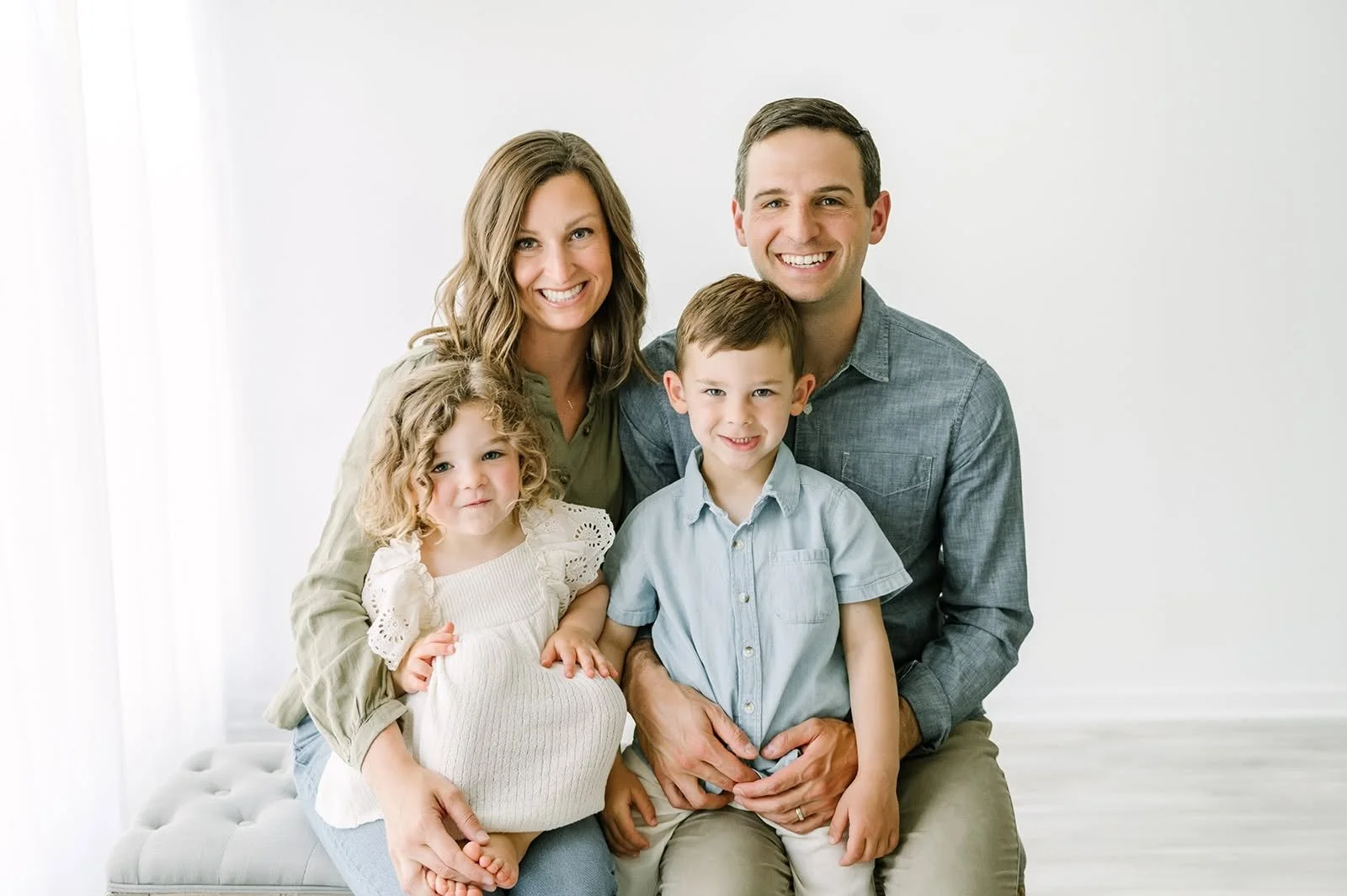 A happy family of four, including a mother, father, and two young children, sitting closely together in a bright, minimalistic room with white walls.