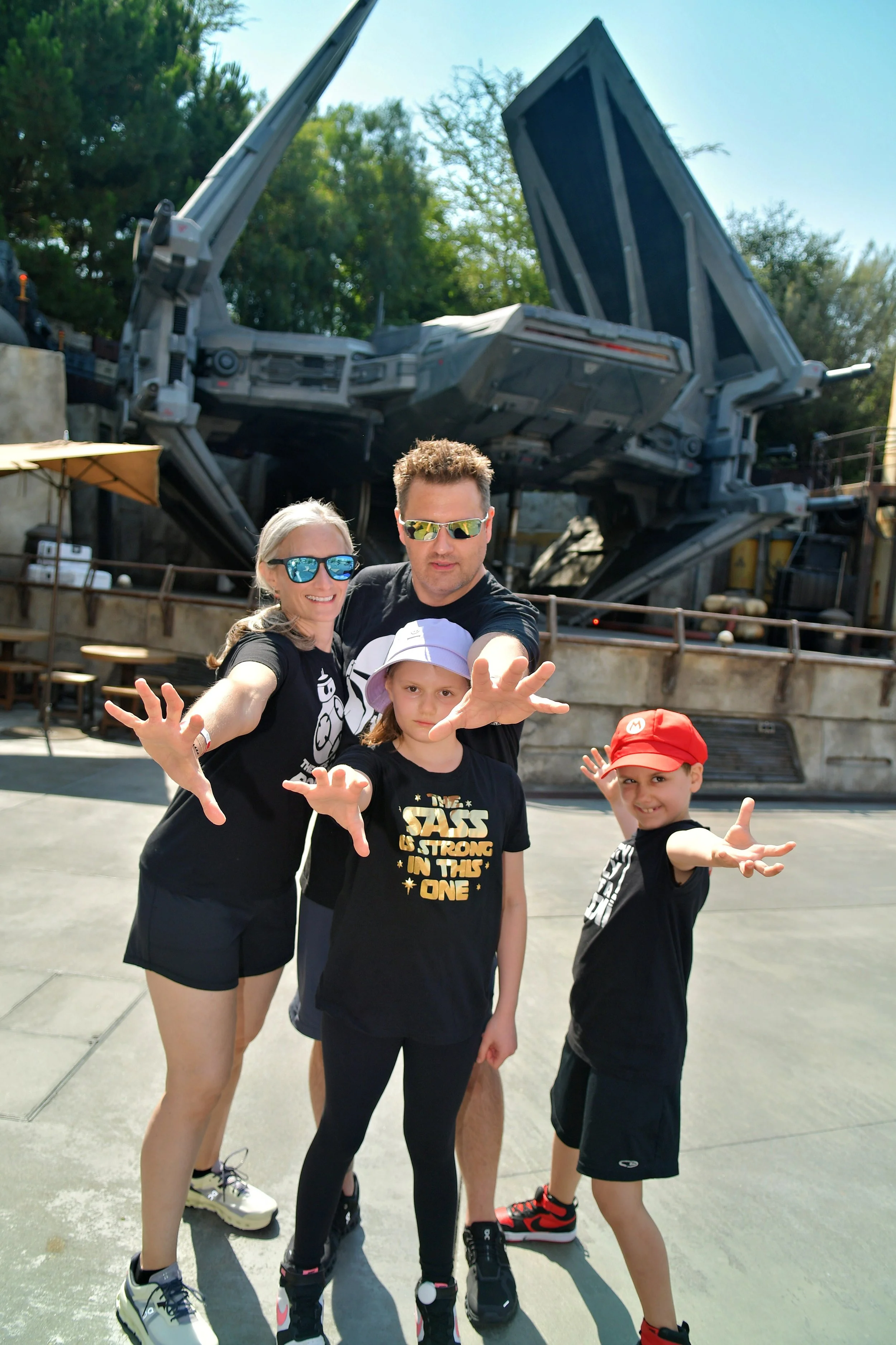 A group of four children and two adults posing with the Star Wars Millennium Falcon ride in the background at a theme park. The children and adults are making hand gestures and smiling.