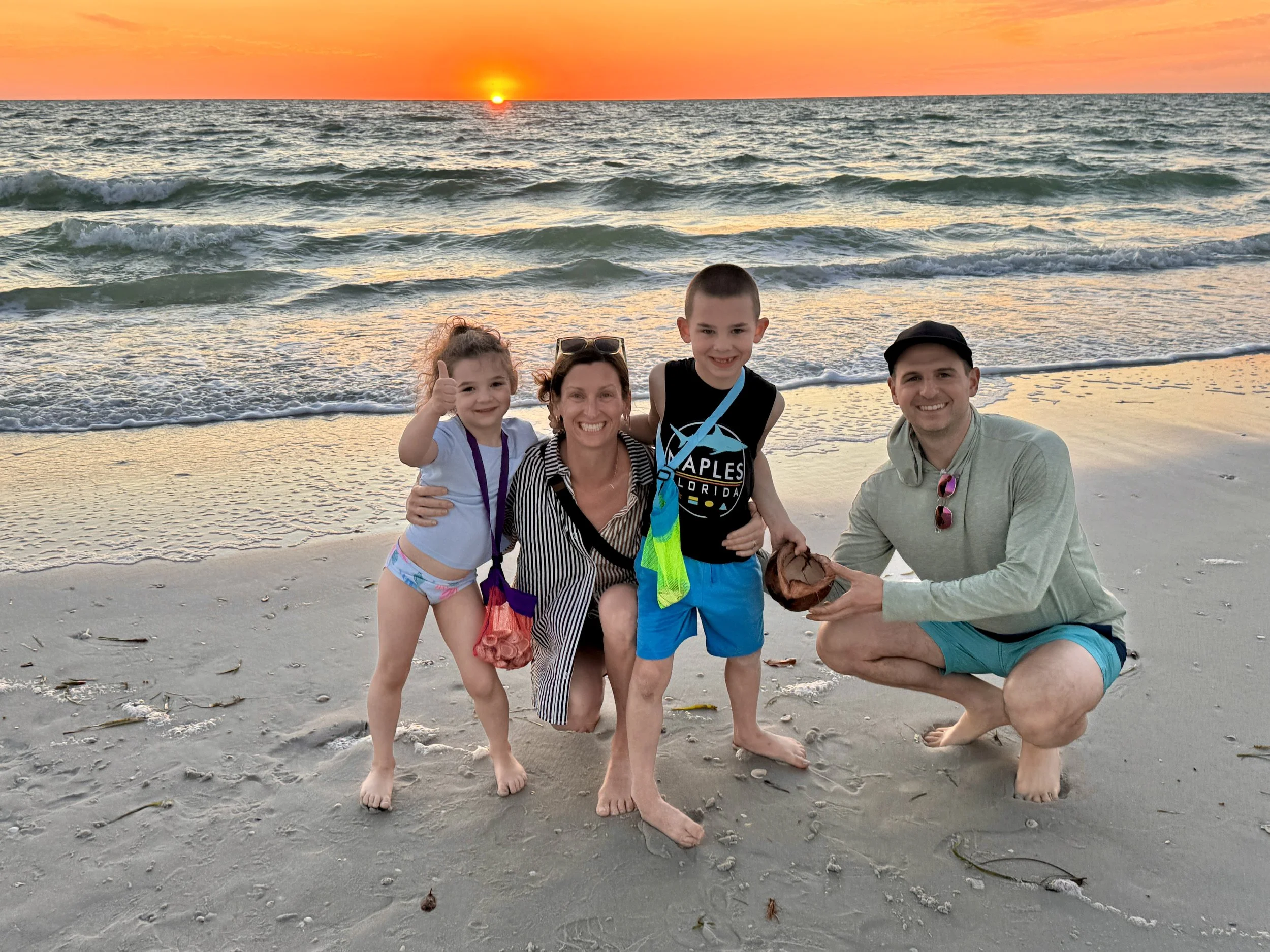 A family of four on the beach during sunset, with the ocean and colorful sky in the background. The family is smiling, with two children and two adults, some kneeling and some standing, near the water's edge.