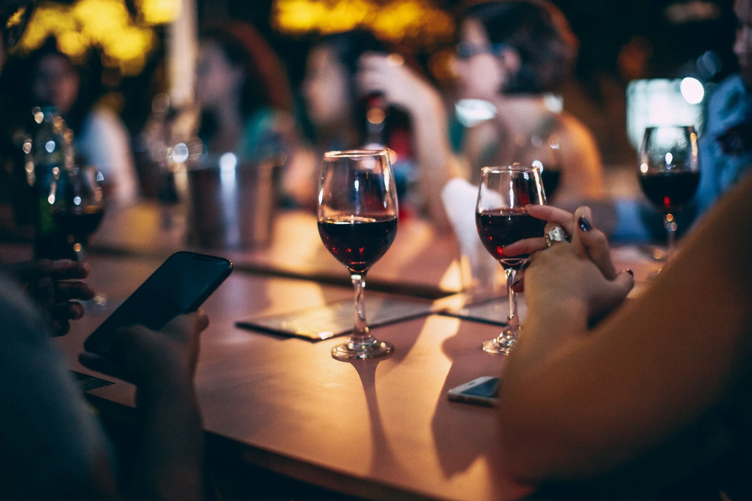 People at a social gathering holding glasses of red wine, with blurted background lights and a table with phones and papers.