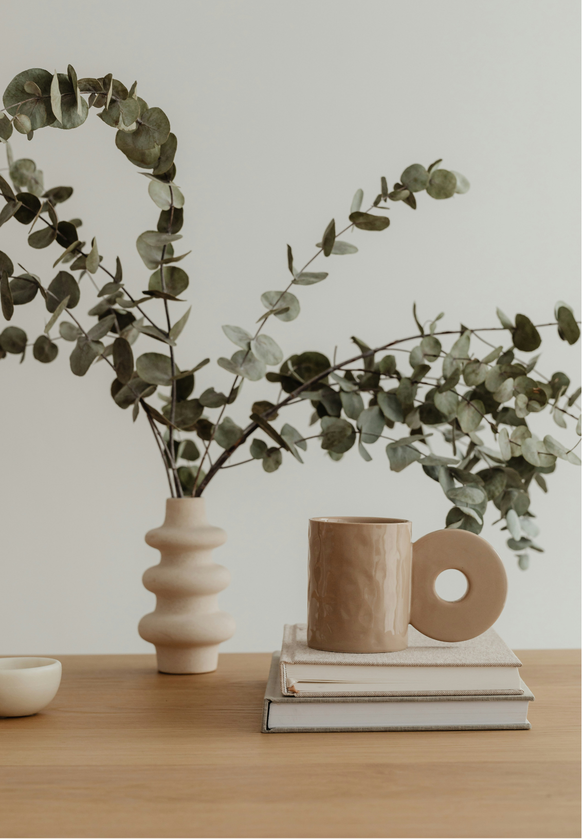 A beige ceramic mug with a round handle sitting on top of two stacked books on a wooden surface, with a tall beige vase holding eucalyptus branches in the background.
