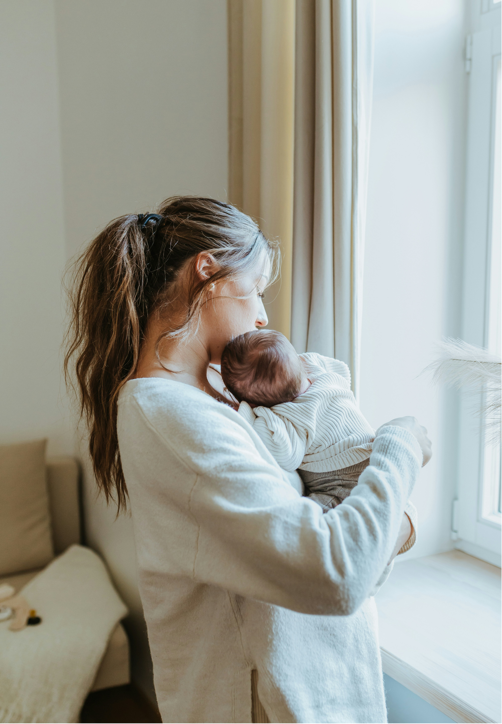 A woman holding a newborn baby near a window with beige curtains, in a softly lit room.