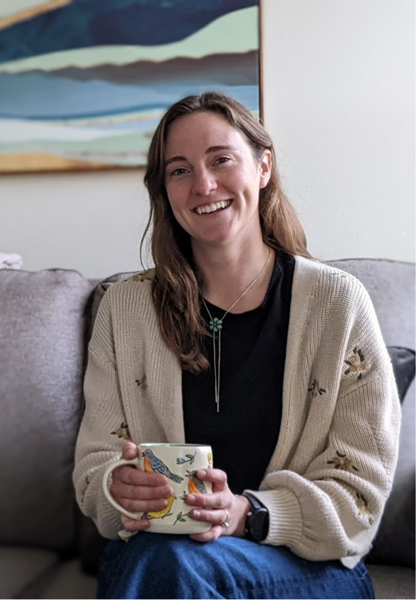 Melissa Ceren, therapist in Colorado Springs, with long brown hair, wearing a beige cardigan, black shirt, and a turquoise flower pendant necklace, sitting on a sofa and holding a mug, in a room with a colorful abstract painting behind her.