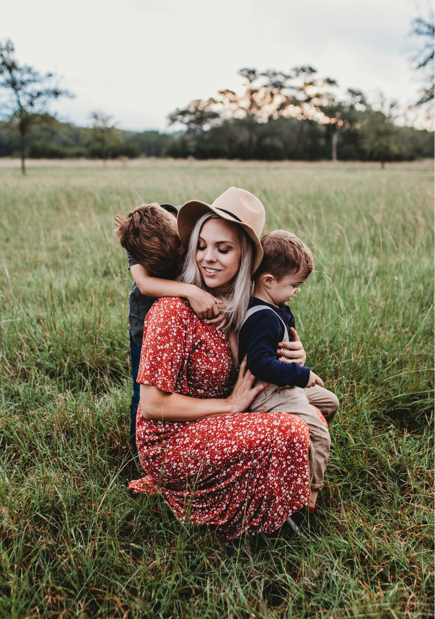 A woman sitting on the grass in a field, holding two young boys who are hugging her from each side. The woman is wearing a wide-brimmed hat and a red floral dress, and the boys are dressed casually. The scene is outdoors with trees in the background and a soft evening sky.