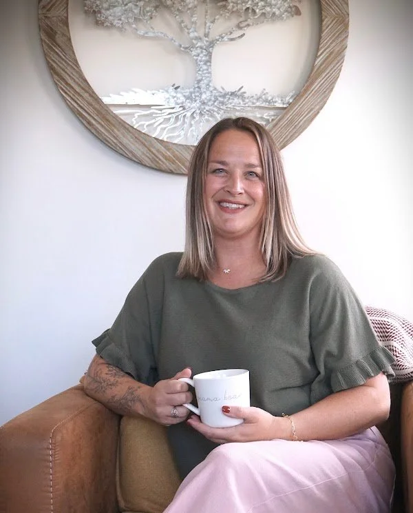Kati Saul, therapist in Colorado Springs, sitting on a sofa holding a white mug, smiling, with a decorative tree wall art behind her.