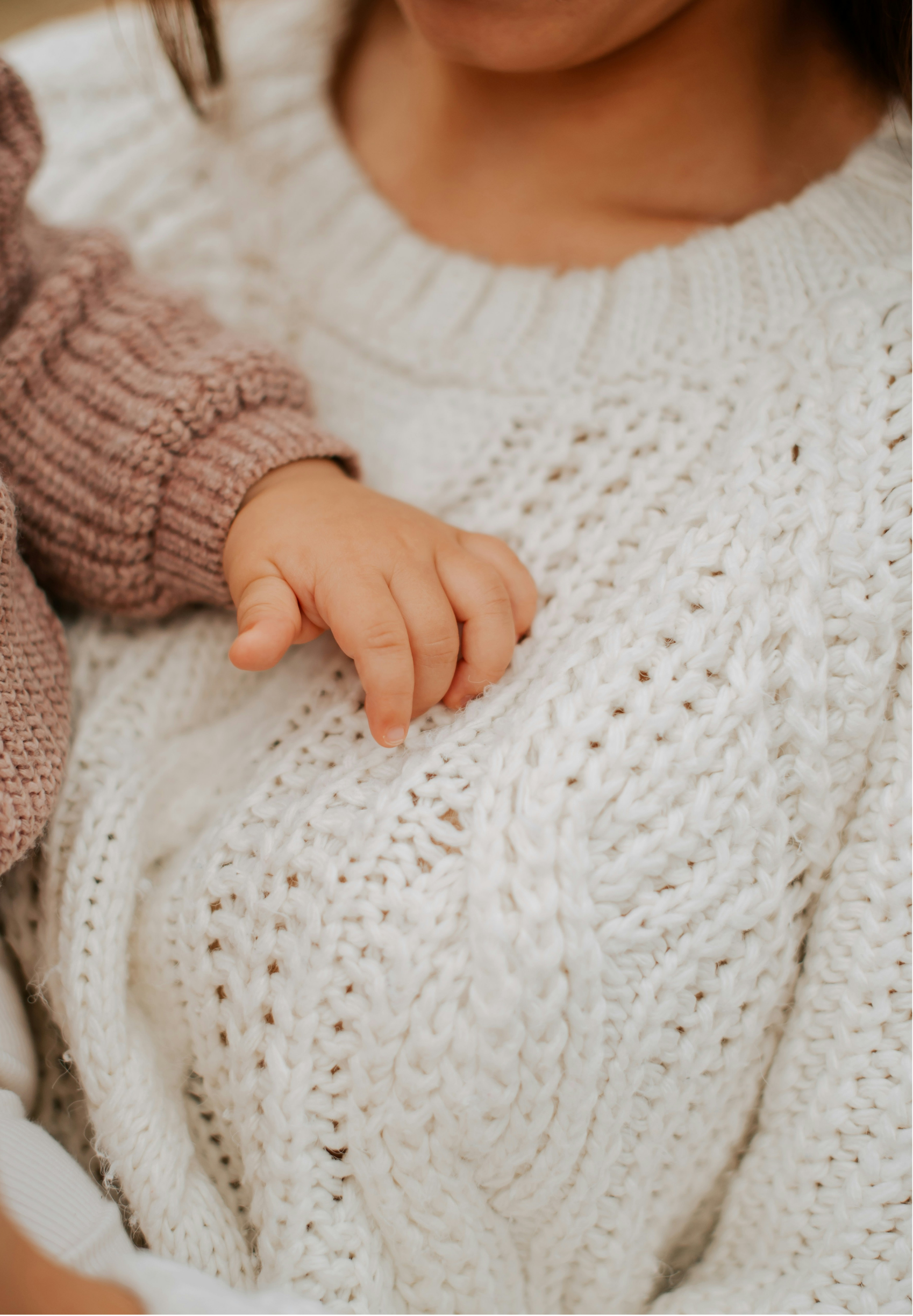 Close-up of a person's hand resting on a chunky white knit blanket, with the person's face partially visible wearing a cream-colored sweater.