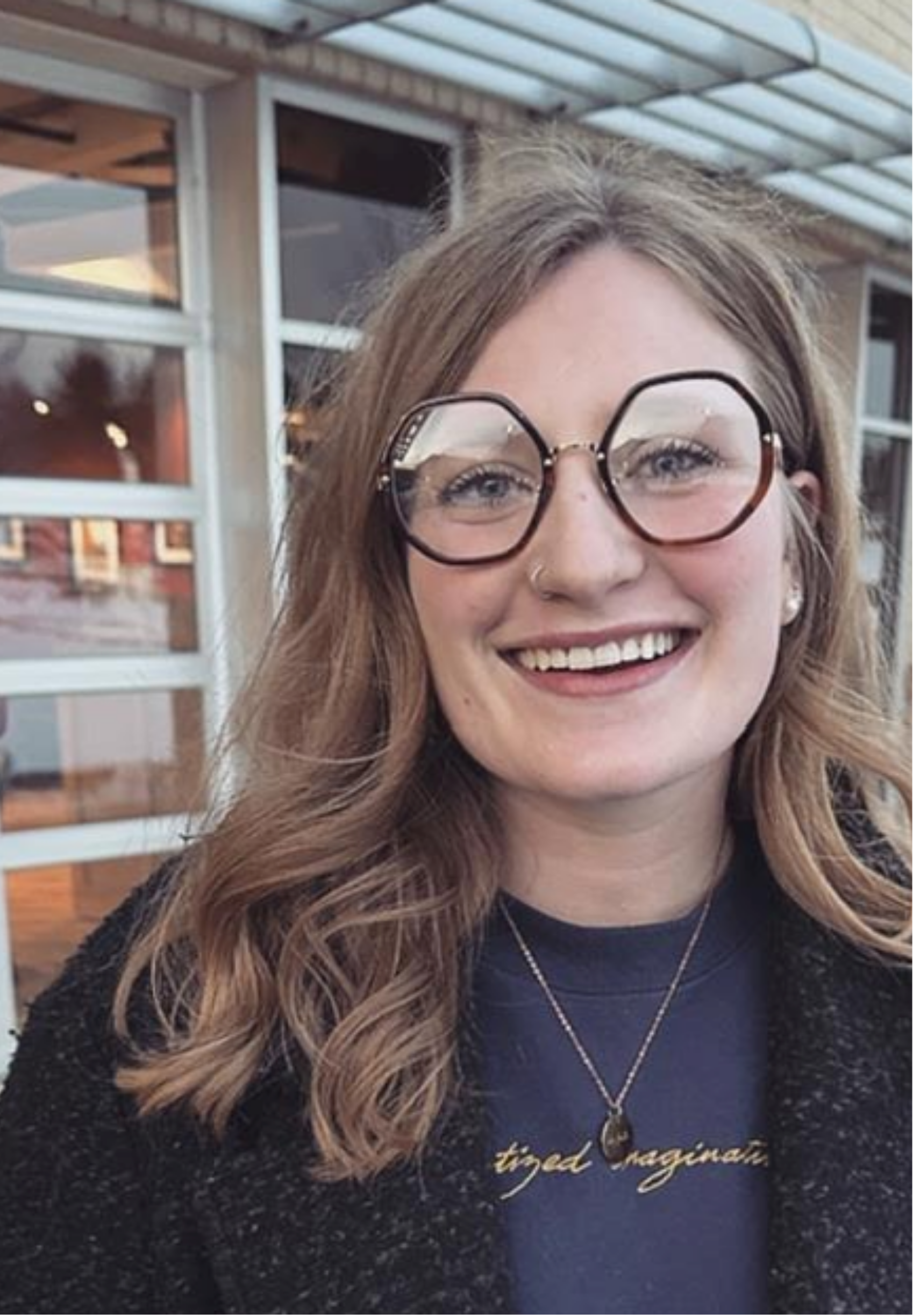 Martha Haller, with long curly hair, glasses, and a nose piercing smiling outdoors near a building with large windows.