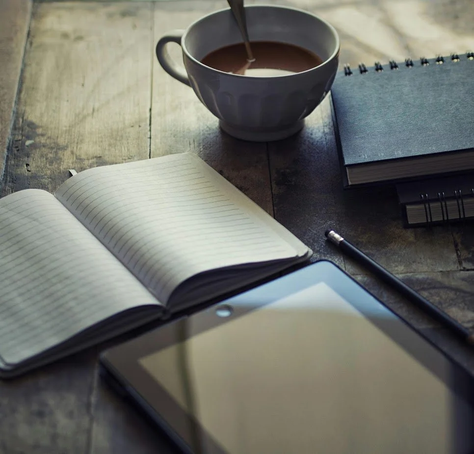 A wooden desk with an open notebook, a cup of coffee, a tablet, a pen, and closed notebooks.