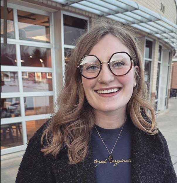 Martha Haller, with wavy, shoulder-length hair and large glasses smiling outdoors, standing in front of a storefront with large windows.