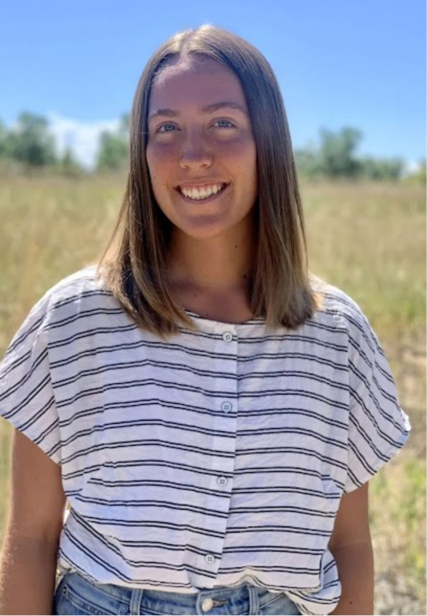Anna Palombi, with shoulder-length brown hair, blue eyes, smiling outdoors under a bright blue sky. She is wearing a white and black striped short-sleeve shirt and high-waisted jeans. The background features a grassy field with some trees.